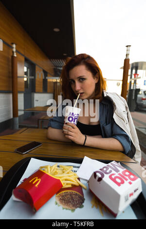 MARUPE LETTONIE - avril 22, 2019 : Jeune femme à boire le café à l'extérieur dans un McDonalds pendant le coucher du soleil sur le terrain Banque D'Images