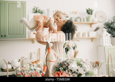 Heureuse mère joue et embrasse son bébé dans la cuisine sur fond de décorations de Pâques. Banque D'Images