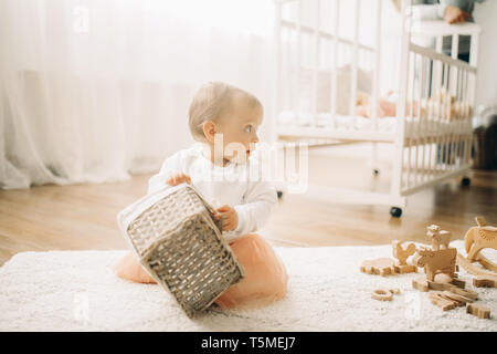 Bébé fille s'assied sur le tapis parmi les jouets en bois et joue avec panier dans la pépinière. Banque D'Images