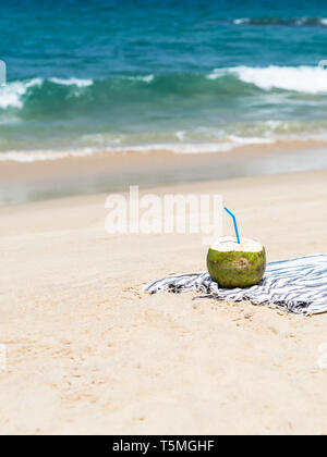 Un nouveau jeune noix de coco est prêt à manger sur une serviette sur une plage de sable fin contre une mer bleue. Vacances tropicales travel concept. Banque D'Images