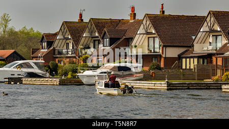 Un seul homme dans un chapeau blanc remontant la rivière Bure dans Horning dans un petit bateau à moteur sur un blanc lumineux et ensoleillé lundi férié. À sa droite Banque D'Images