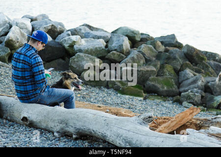 Un homme adulte en costume bleu occasionnels examine son téléphone pendant que son chien se trouve sur la plage de Myrtle Edwards Park à Seattle aux Etats-Unis. Banque D'Images