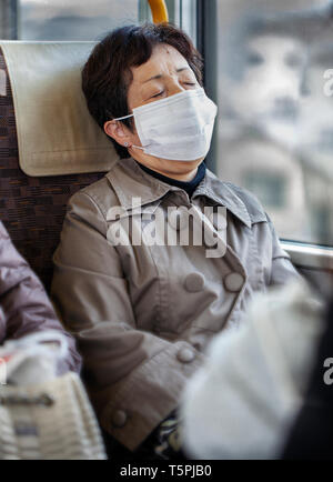 Older Asian woman wearing a tan de l'imperméable et chirurgicaux masque pollution assis sur un bus train Metro Banque D'Images