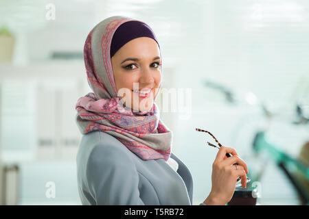 Belle femme élégante en hijab et lunettes, sitting at desk with laptop in office. Portrait of businesswoman musulmane. Bureau moderne avec Banque D'Images