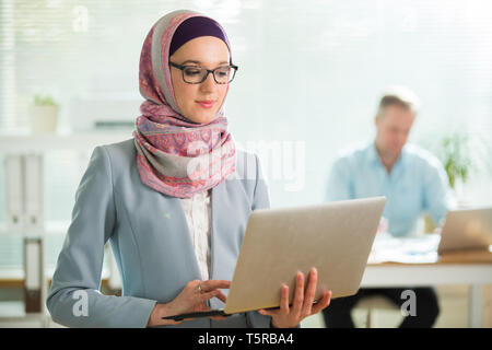 Belle femme élégante en hijab et lunettes standing in office, holding laptop. Portrait of businesswoman musulmane. Bureau moderne avec de grandes Banque D'Images