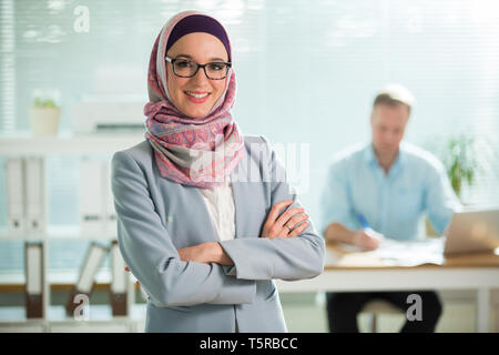Belle jeune femme qui travaille à l'hijab, costume et lunettes standing in office, en souriant. Portrait of businesswoman musulmane. Wit bureau moderne Banque D'Images