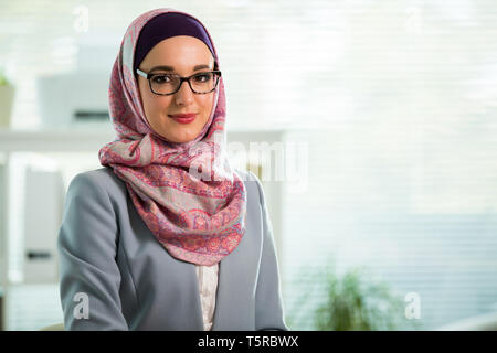 Belle femme qui travaille à l'hijab et eyeglasses smiling in office, holding folders et ordinateur portable dans les bras. Portrait of businesswoman musulmane Banque D'Images