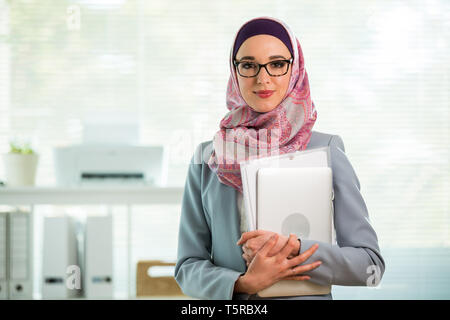 Belle femme qui travaille à l'hijab et eyeglasses smiling in office, holding folders et ordinateur portable dans les bras. Portrait of businesswoman musulmane Banque D'Images