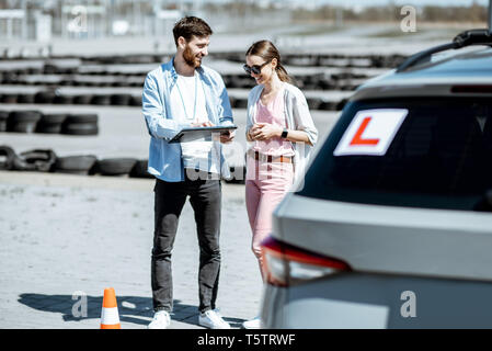 Instructeur mâle montrant plans routiers pour une étudiante, debout près de la voiture d'apprentissage sur le terrain d'entraînement en plein air Banque D'Images
