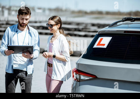 Instructeur mâle montrant plans routiers pour une étudiante, debout près de la voiture d'apprentissage sur le terrain d'entraînement en plein air Banque D'Images