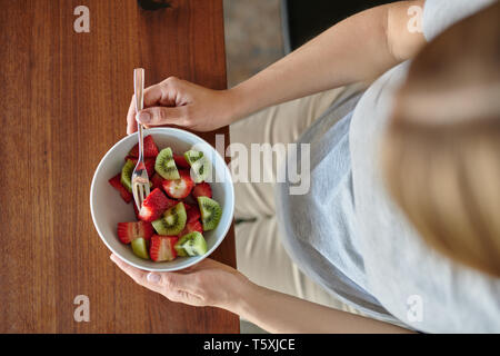 Pregnant woman eating un bol de salade de fruits avec des fraises fraîches assis à une table à manger dans un gros plan sur l'aliment et son ventre dans un régime alimentaire sain Banque D'Images