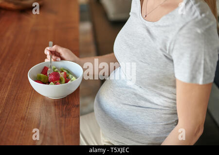 Pregnant woman eating un bol de salade de fruits avec des fraises fraîches assis à une table à manger dans un gros plan sur l'aliment et son ventre dans un régime alimentaire sain Banque D'Images