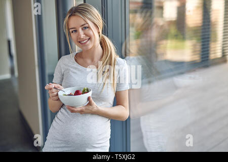 Heureux femmes enceintes en bonne santé jeune blonde femme debout appuyé contre une fenêtre patio smiling at the camera tout en appréciant un bol de salade de fruits frais Banque D'Images