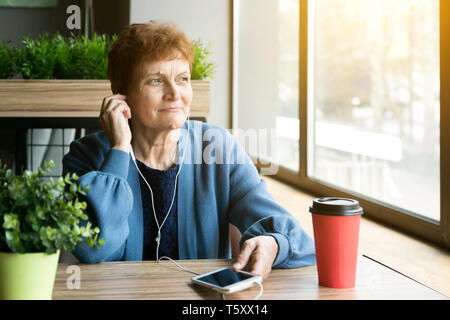 Pensionné mature assis dans un café à boire du café et à l'écoute de chansons préférées dans le casque du téléphone. Banque D'Images