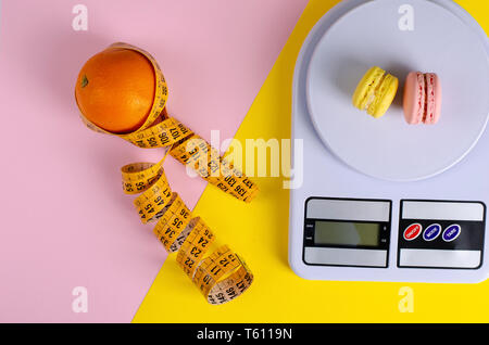 Orange avec un ruban à mesurer, balance de cuisine numérique avec ses macarons sur fond jaune et rose. Équilibre entre l'alimentation et la mauvaise alimentation. Vue d'en haut Banque D'Images