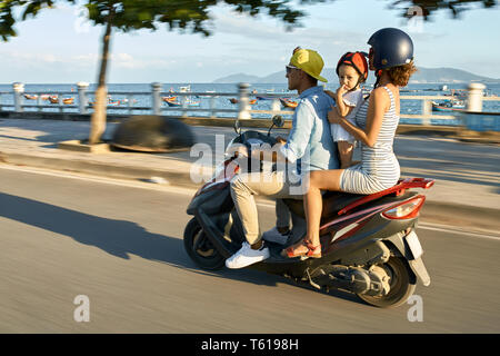 Joli couple avec une petite fille sont équitation moto rouge sur la ville rue sur fond de mer baie ensoleillée avec de nombreux bateaux au Vietnam. Ils wearin Banque D'Images