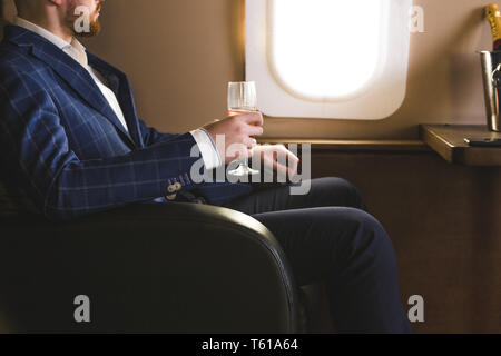 Un jeune homme dans un costume coûteux est assis dans le fauteuil d'un jet privé avec un verre de champagne dans sa main et regarde par la fenêtre Banque D'Images