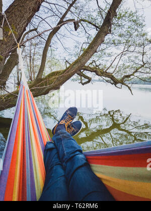 Homme avec chaussures bleu jeans et balançoires dans hamac coloré s'interrompt sur le bord du lac au Danemark Banque D'Images