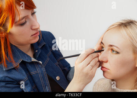 Maquilleur aux cheveux rouges impose le maquillage sur les yeux fermés à l'aide d'un pinceau applique des ombres sur les paupières dans un style oriental : Golden, marron et vert Banque D'Images