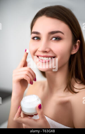 Smiling young woman applying face cream sur sa peau parfaite. Modèle avec la peau impeccable holding white pot de crème. Banque D'Images