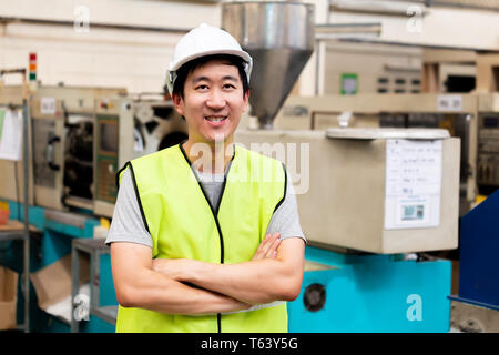 Vue avant du travailleur d'usine asiatique avec casque de sécurité posés à la caméra avec au sourire heureux dans les installations industrielles à l'usine de l'industrie lourde Banque D'Images
