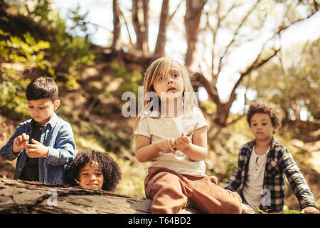 Cute girl sitting on a log avec des bâtons dans la main et ses amis autour de la région de forêt. Garçons et une fille jouant dans la forêt. Banque D'Images