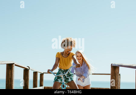Deux femmes amis jouant avec un vélo sur la promenade le long de la mer. Femme assise sur le vélo avec ses amis poussant par l'arrière et souriant. Banque D'Images
