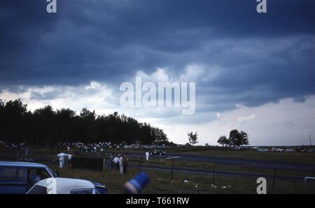 Les spectateurs, sur une journée nuageuse, regarder une course automobile à une version de l'hippodrome international de Watkins Glen, avec les gens et les voitures garées dans les champs environnants, Watkins Glen, New York, 1965. () Banque D'Images