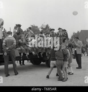 1967, historique, groupe de jeunes garçons habillés comme des cowboys et des Indiens s'amusant sur l'arrière d'un camion à un carnaval ou fête de village, entre 2 lacs en dollars, en Angleterre. Banque D'Images