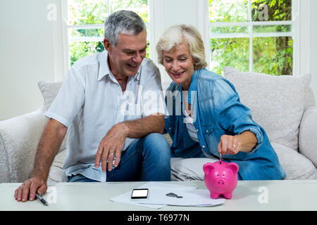 Senior woman putting coins in piggy bank lors d'un entretien avec l'homme sur canapé Banque D'Images