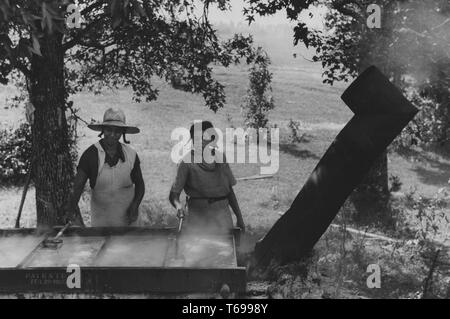 Photographie en noir et blanc de deux femmes afro-américaines, l'un portant un floppy hat, un tablier, et un léger sourire sur son visage, l'autre portant une robe d'impression, à la fois à la tonnellerie de rectiligne en direction de Simmering jus de canne à sucre qu'ils écumer avec tamis à long manche ; situé à Orange County, North Carolina, USA ; photographié par Marion Poster Walcott, sous le parrainage des membres de l'Administration de la sécurité agricole, septembre 1939. À partir de la Bibliothèque publique de New York. () Banque D'Images