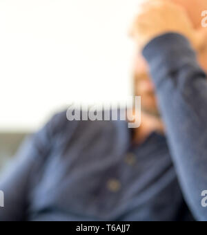 Épuisé homme d'âge moyen avec une barbe est assis sur un canapé et repose sa tête fatiguée dans sa main defocused Banque D'Images