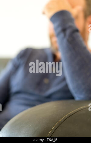 Épuisé homme d'âge moyen avec une barbe est assis sur un canapé et repose sa tête fatiguée dans sa main focus sélectif Banque D'Images