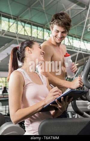 Trainer écrit sur un presse-papiers tandis que l'homme exerce sur une machine elliptique Banque D'Images