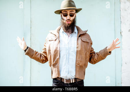 Portrait d'un homme barbu élégant habillé en veste et chapeau sur la lumière fond bleu turquoise à l'extérieur Banque D'Images