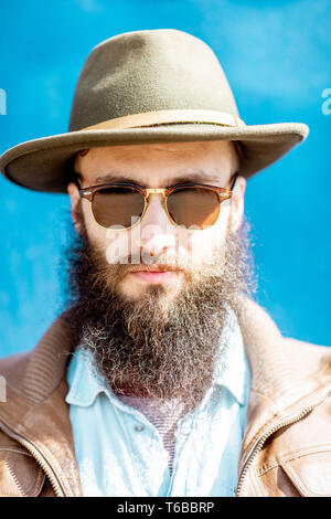 Close-up portrait d'un homme barbu élégant avec chapeau et lunettes de soleil sur le mur bleu Banque D'Images