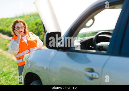 Une femme en veste orange Ouvrir capot de voiture voiture cassée - penser la façon de résoudre le problème Banque D'Images