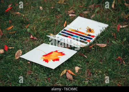 Peinture de jaune rouge l'automne feuille d'érable sur du papier blanc et fort des peintures utilisées allongé sur l'herbe verte entre les feuilles d'automne. Still Life de hobby Banque D'Images