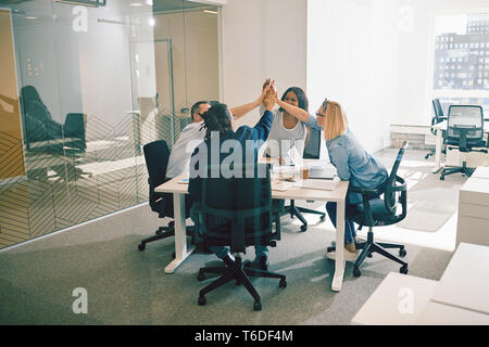 Groupe diversifié de collègues de travail de sourire et de plafonnement élevé ensemble au cours d'une réunion autour d'une table dans un bureau moderne et lumineux Banque D'Images