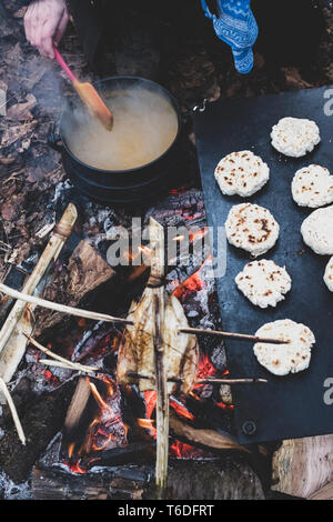 High angle close up de pains fraîchement préparés et de poisson sur les brochettes sur un feu de camp, et une personne en remuant la soupe dans le pot de fer de fonte. Banque D'Images