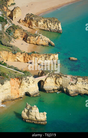 VUE AÉRIENNE. Série de falaises parallèles en bord de mer à Praia dos Estudantes (plage des étudiants), pont romain en premier plan. Lagos, Algarve, Portugal. Banque D'Images