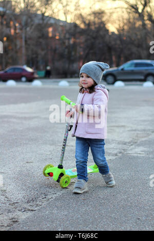 Cute girl dans un chapeau, manteau et jeans équitation un scooter à trois roues. La ville de printemps soirée coucher du soleil. Banque D'Images