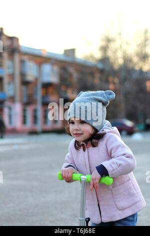 Cute girl dans un chapeau, manteau et jeans équitation un scooter à trois roues. La ville de printemps soirée coucher du soleil. Banque D'Images