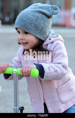 Cute girl dans un chapeau, manteau et jeans équitation un scooter à trois roues. La ville de printemps soirée coucher du soleil. Banque D'Images