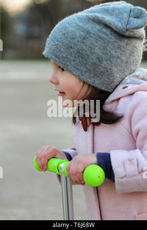 Cute girl dans un chapeau, manteau et jeans équitation un scooter à trois roues. La ville de printemps soirée coucher du soleil. Banque D'Images