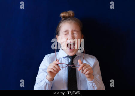 Fatigué d'écolière élémentaire de bâiller. Épuisé little girl holding lunettes. Enfant paresseux dans l'uniforme scolaire sur fond bleu foncé. Enfant endormi, leçon ennuyeuse, étudiant surmené Banque D'Images