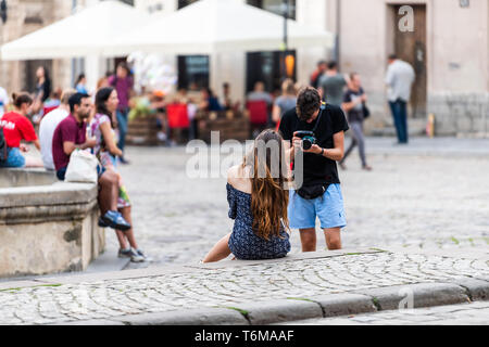 Lviv, Ukraine - le 30 juillet 2018 : la ville de Lvov polonais ukrainien historique durant la journée avec fille assise sur le banc et petit ami de prendre photo d'elle avec sont venus Banque D'Images