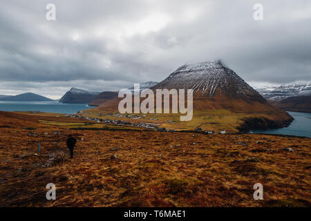 Randonneur solitaire en route vers le haut de la Kap Enniberg avec vue de Viðareiði et son fjord avec des montagnes neige-couvertes (îles Féroé, Danemark, Europe) Banque D'Images
