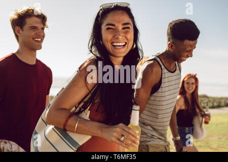 Cheerful young woman holding une bouteille de bière la marche à l'extérieur avec des amis. Amis aller à la plage avec un verre un jour d'été. Banque D'Images