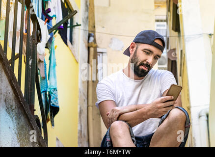 Portrait d'un homme avec une barbe, style décontracté, l'envoi de messages - bavardant avec son smartphone mobile, assis dans la rue, à l'extérieur. Banque D'Images